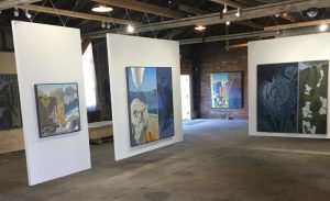 Paintings displayed on white partitions in an industrial gallery space with exposed brick walls.
